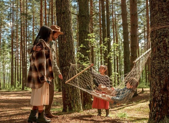 A family relaxes in a hammock amidst tall trees, enjoying a sunny day outdoors.