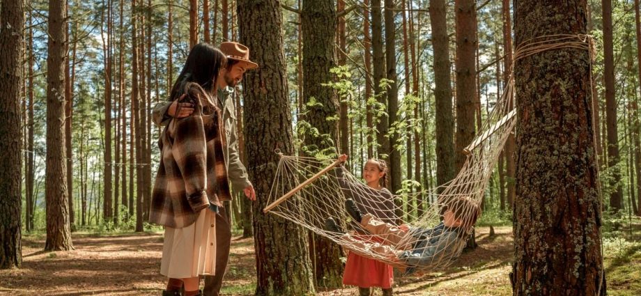 A family relaxes in a hammock amidst tall trees, enjoying a sunny day outdoors.